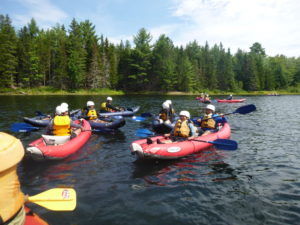 Group of kids sitting in red and blue inflatable kayaks taking a break from paddling on a pond