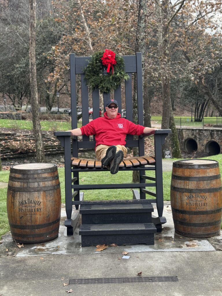 Person sitting in oversized rocking chair in park area at Jack Daniels facility. There is a wreath hanging on the chair and two Jack Daniels barrels on each side of the chair.