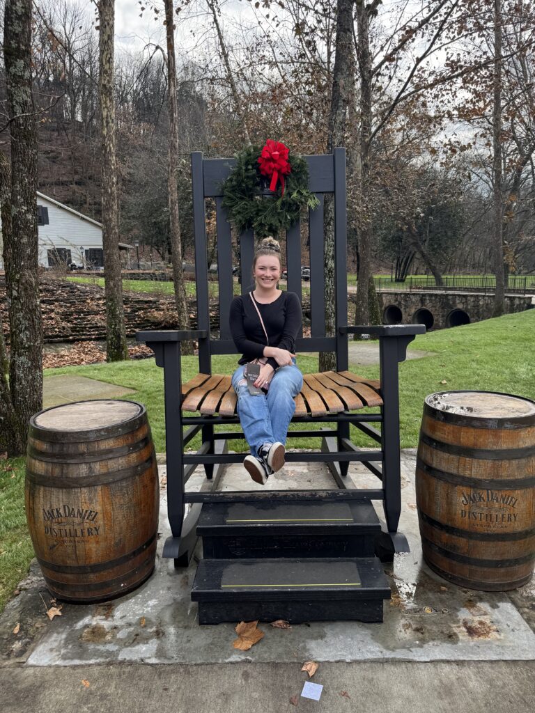 Person sitting in oversized rocking chair in park area at Jack Daniels facility. There is a wreath hanging on the chair and two Jack Daniels barrels on each side of the chair.