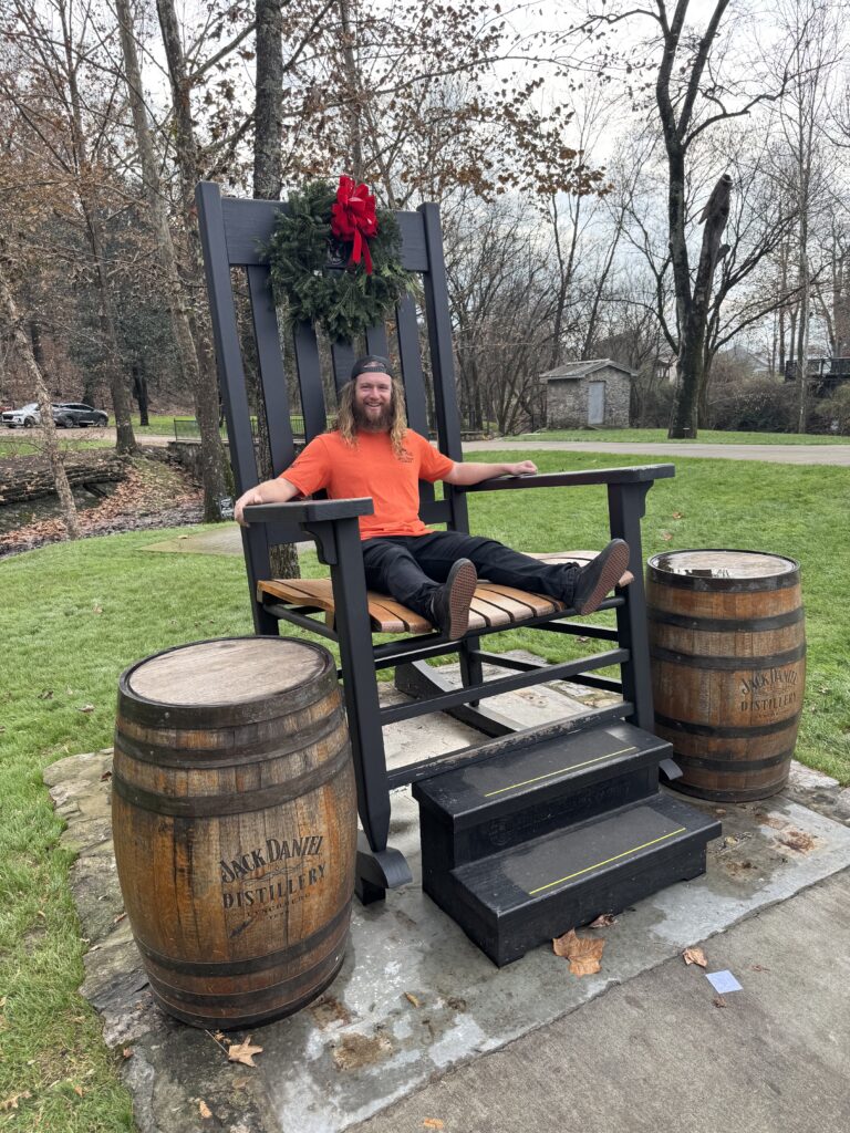 Person sitting in oversized rocking chair in park area at Jack Daniels facility. There is a wreath hanging on the chair and two Jack Daniels barrels on each side of the chair.