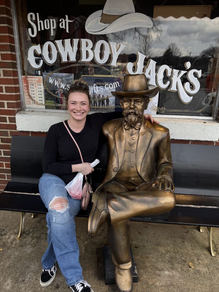 Chastity sitting next to life size gold statue of a man on a bench outside of shop that says "Shop at Cowboy Jacks's" on the shop window behind the bench.