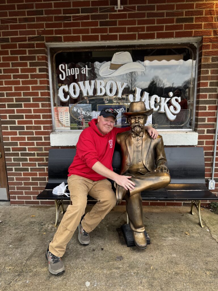 Man in red sweatshirt sitting next to life size gold statue of a man on a bench outside of shop that says "Shop at Cowboy Jacks's" on the shop window behind the bench.