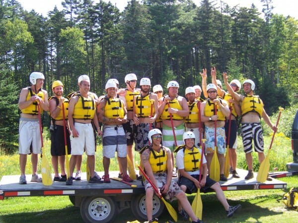 Group of People on a Trailer Smiling With Whitewater Rafting Gear On. All wearing yellow life jackets and white helmets